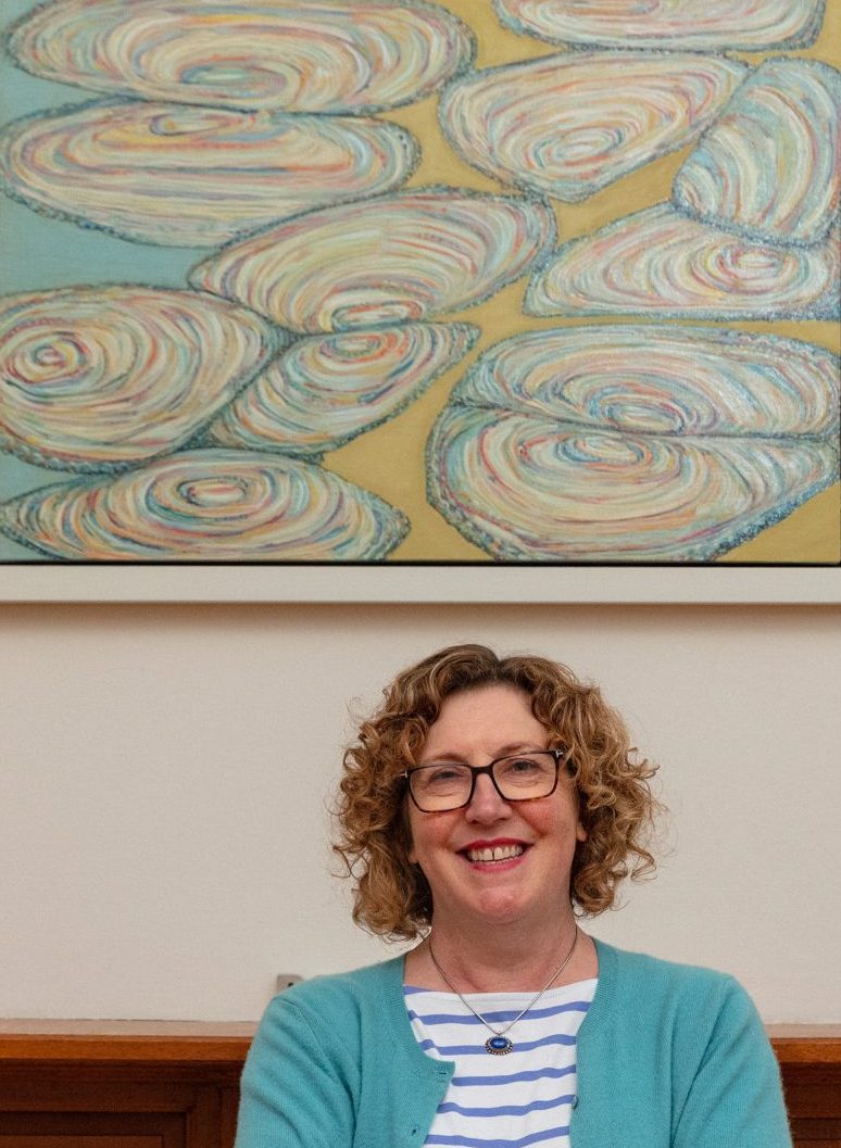 Interior headshot of artist Sarah Kudirka; a white woman with curly fair hair and glasses, wearing a striped top, necklace and turquoise blue cardigan, smiling with face to camera, and a painting of shell-like abstracted forms hung behind her on a pale wall above a wood fireplace. Photographer credit: Alexander Hoyles